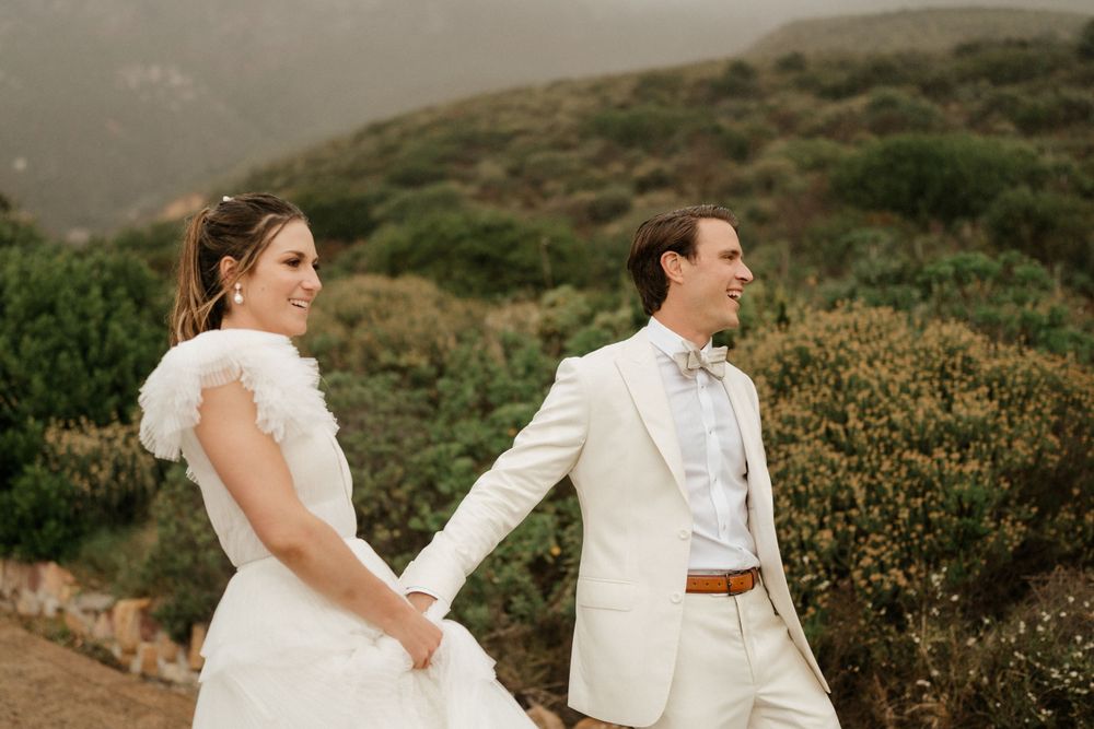 Couple walking hand in hand through coastal scenery in elegant white wedding attire.