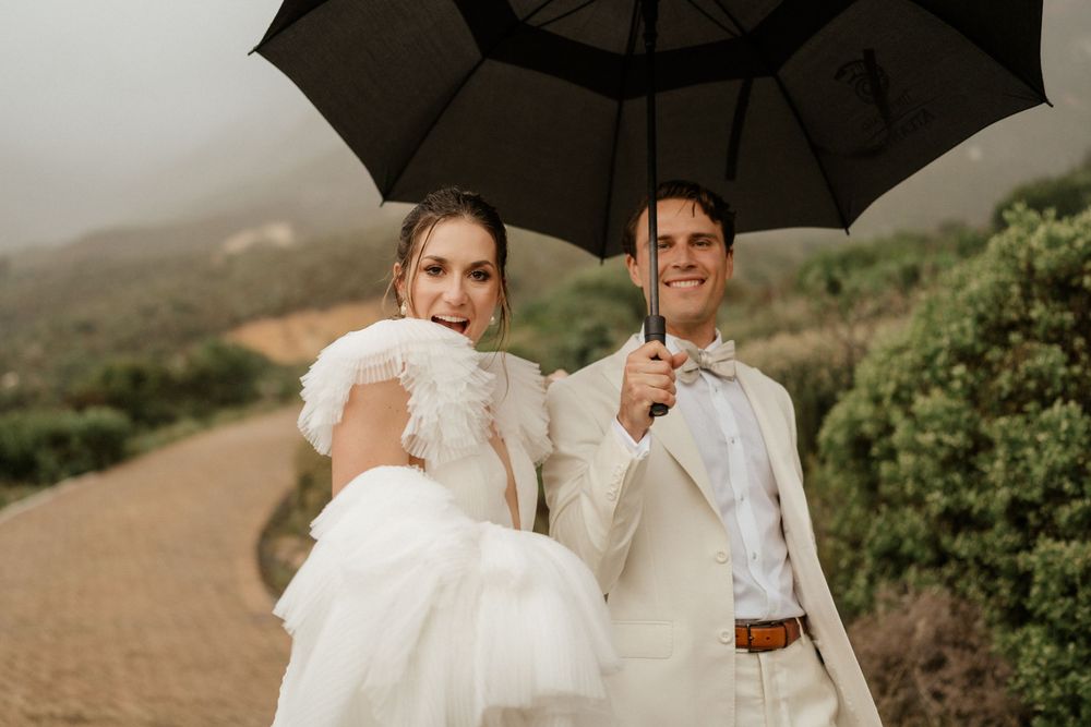 Newlyweds share a romantic moment under a black umbrella on a misty mountain path.