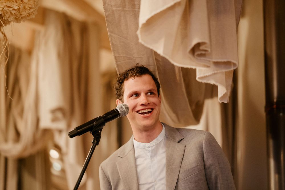 A speaker in a light gray suit delivers a speech at an elegant venue with draped fabric decor.