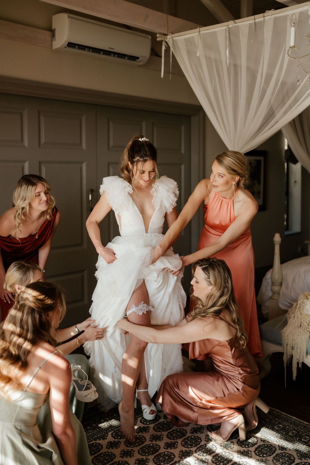 Bridesmaids in coral dresses help adjust the bride's gown during wedding preparations.