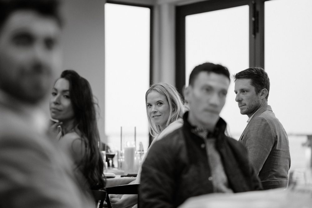 Candid black and white photograph of guests mingling at a social gathering near windows.