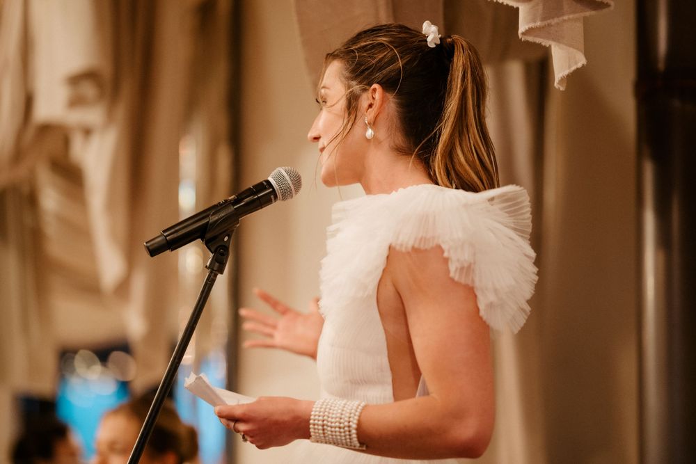 A speaker in a white dress delivers remarks at an indoor wedding reception.