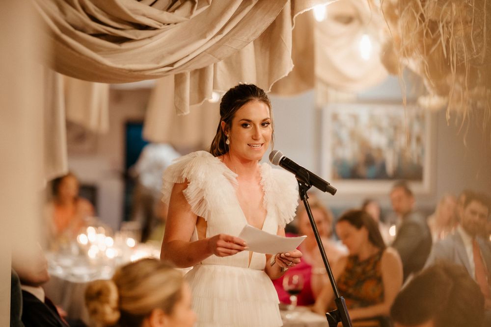 Person in white dress gives speech at wedding reception with decorative draping overhead.