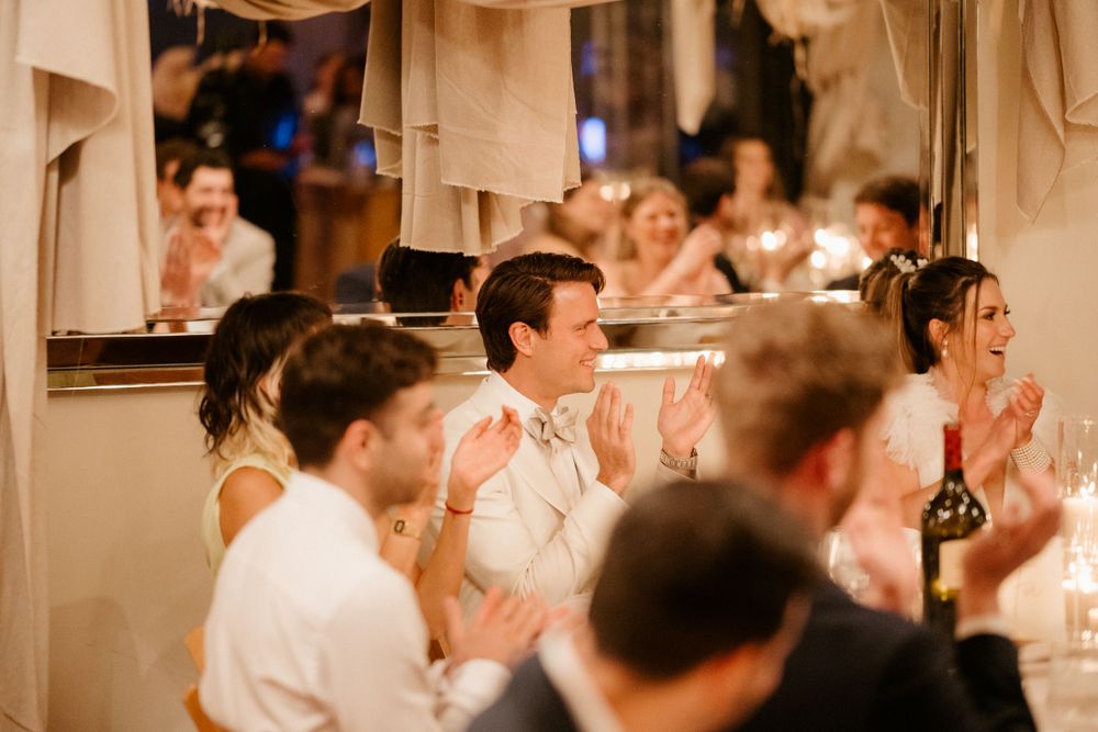Wedding reception guests in white attire clapping during an indoor celebration.