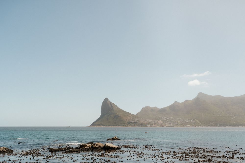 Dramatic coastal landscape with rocky shoreline and mountain peaks against clear blue sky.