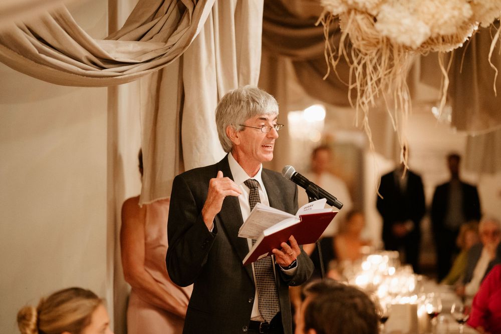 Officiant reads from a book during an indoor wedding ceremony with draped fabric decor and candlelight.