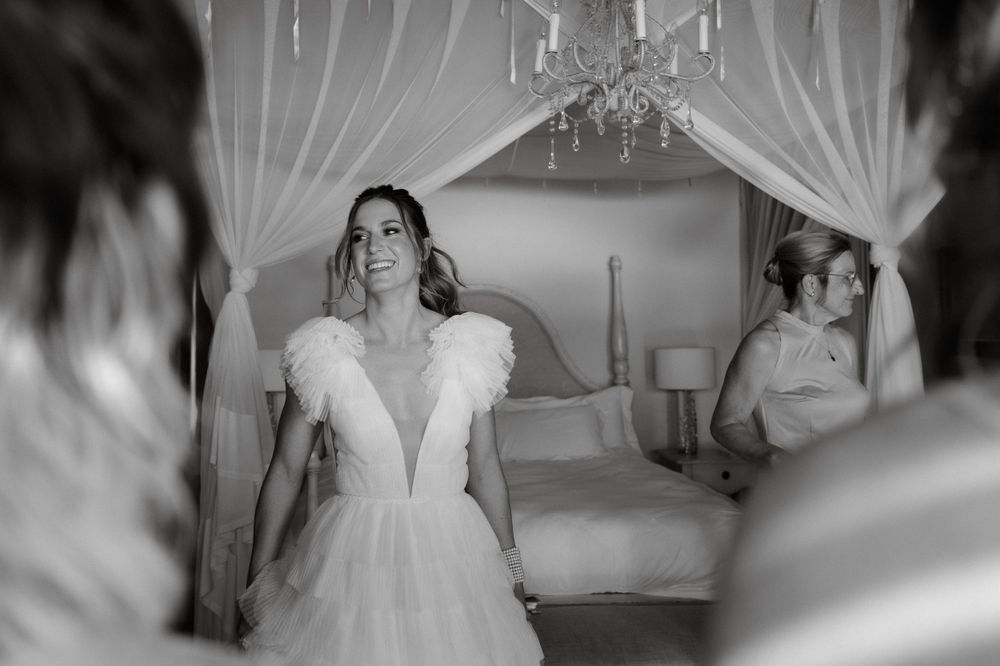 A bride in a white wedding dress with feather details stands in a room with flowing white curtains and a chandelier.