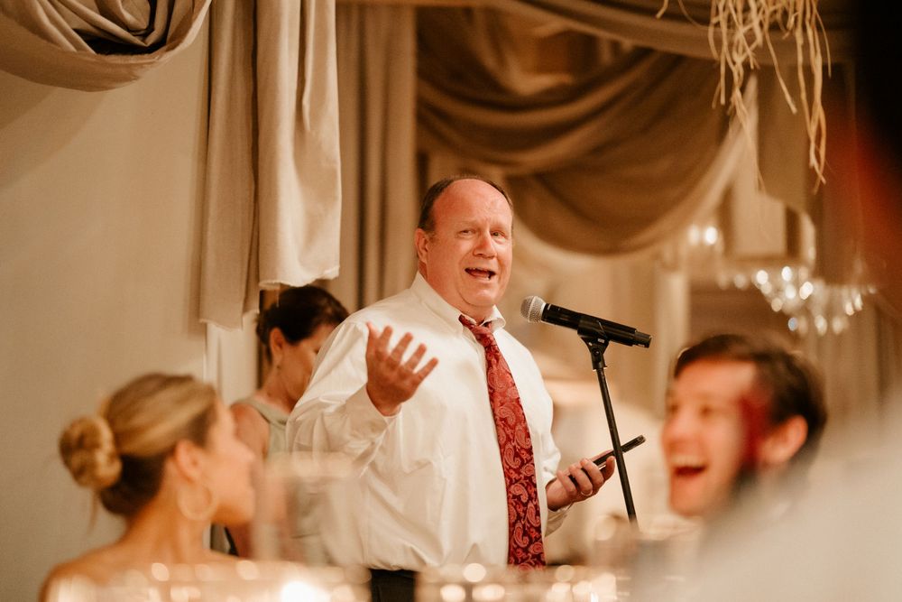 An enthusiastic toast speech being given at an elegant wedding reception with chandelier lighting.