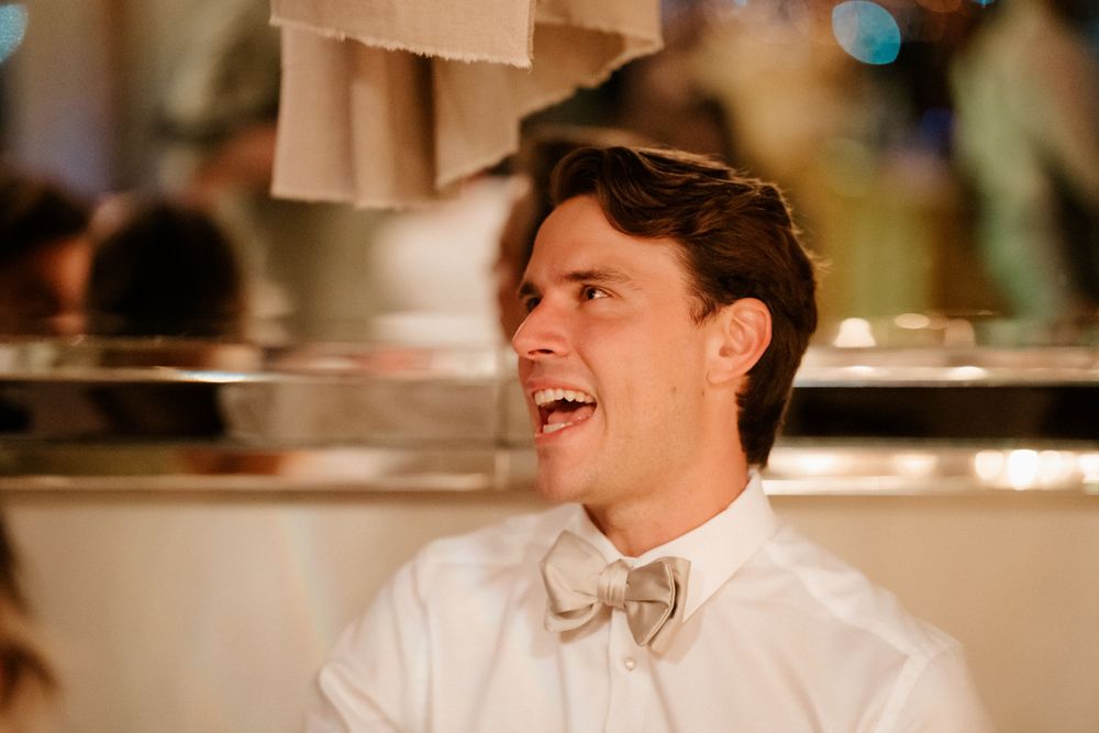 A guest in a white shirt and bowtie laughs joyfully during wedding festivities.