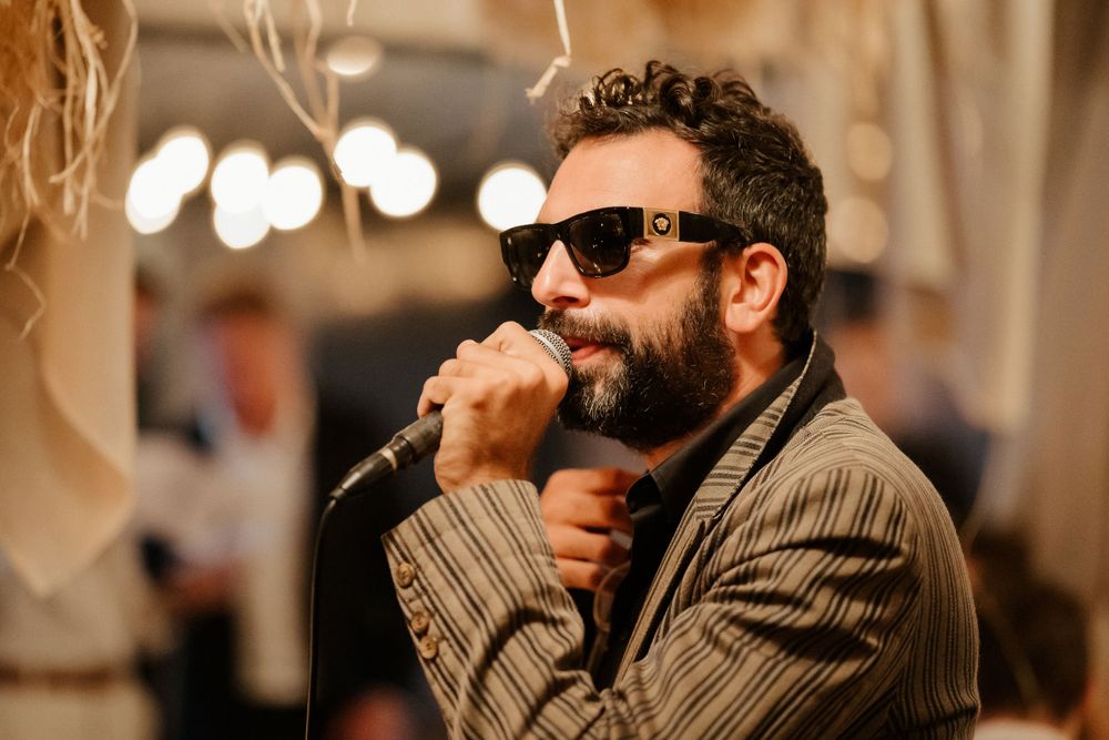 Bearded performer in sunglasses and suit jacket singing emotively into microphone at indoor event.