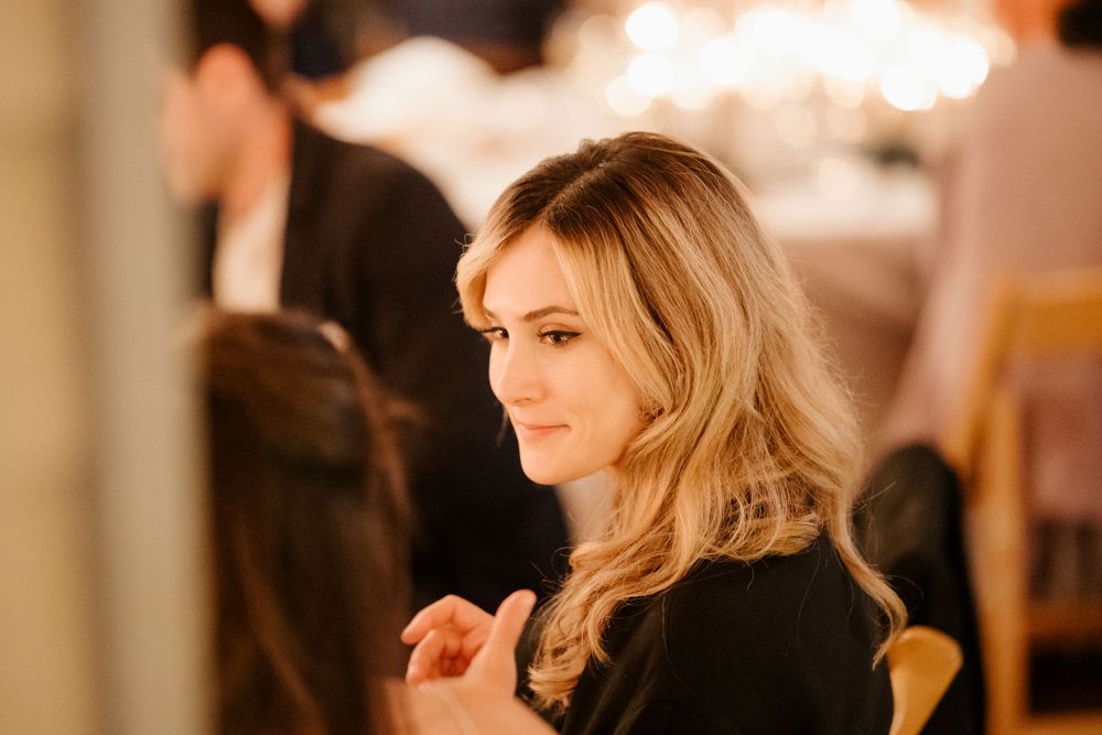 A woman with wavy blonde hair in a black outfit standing in a warmly lit room.