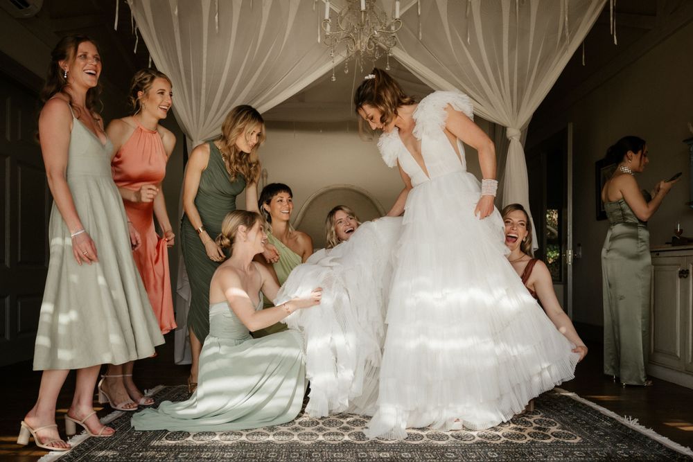 Wedding party helping with dress details during preparation in a dimly lit room with white curtains.