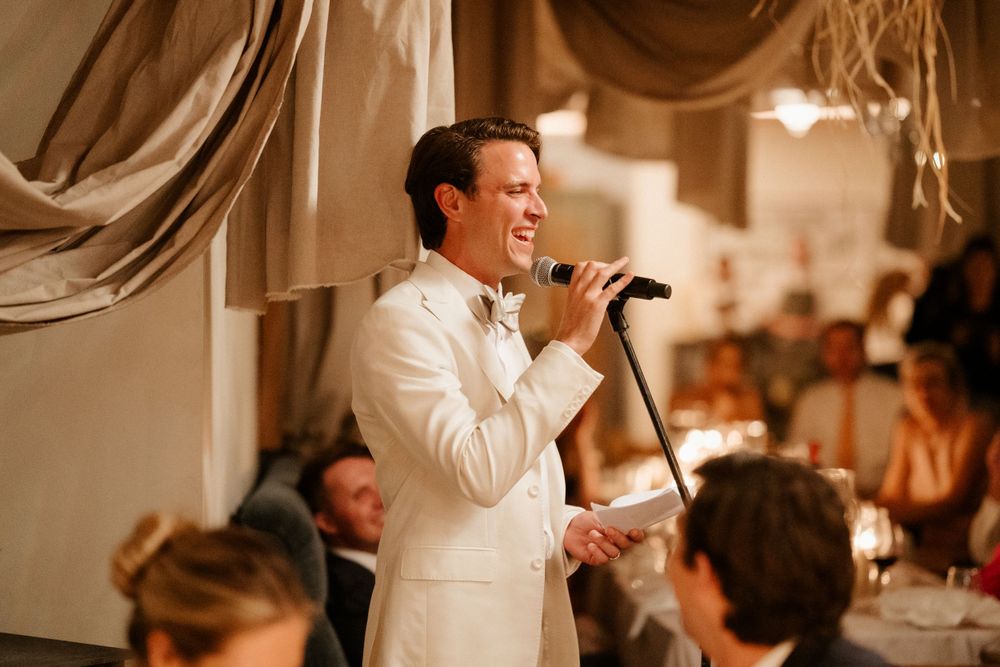 A performer in a white suit jacket holds a microphone while singing at an elegant indoor reception venue.