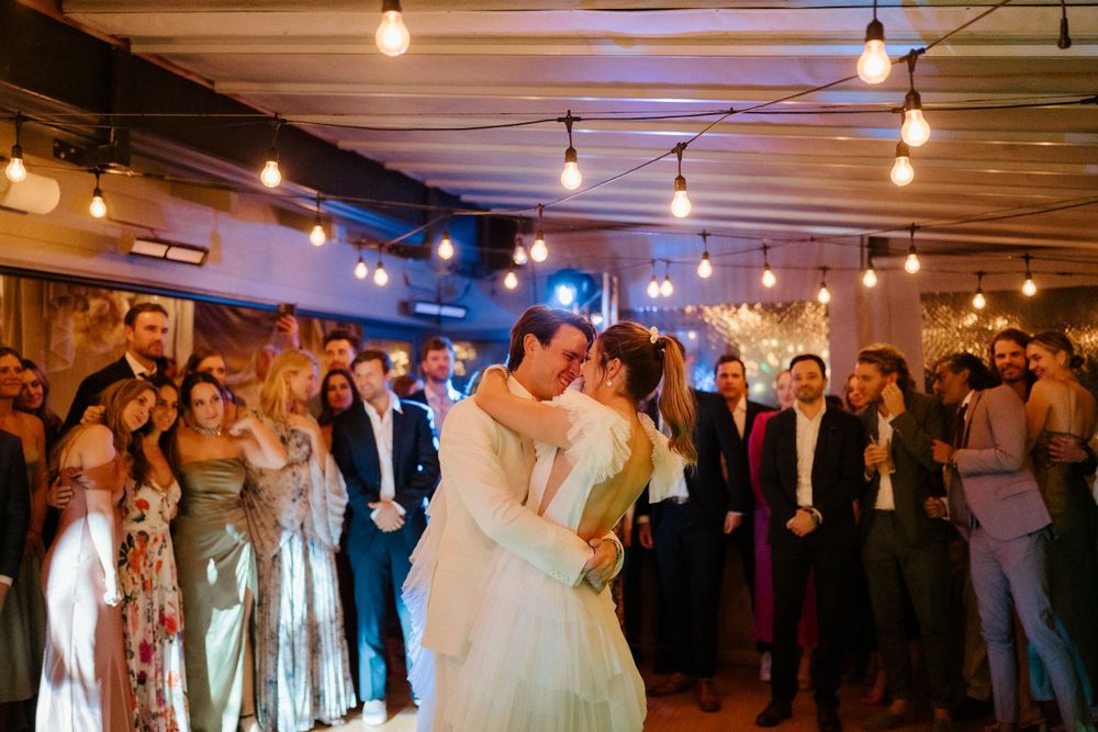 Newlyweds share their first dance surrounded by guests under warm string lights.