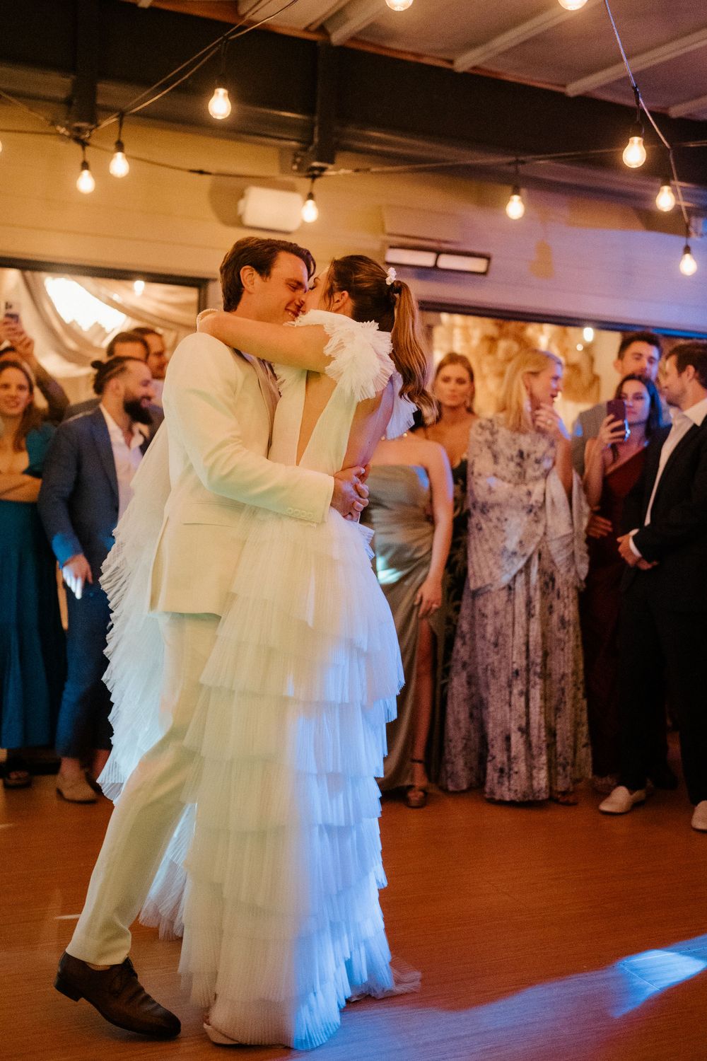 Newlyweds share their first dance surrounded by guests under industrial string lights.