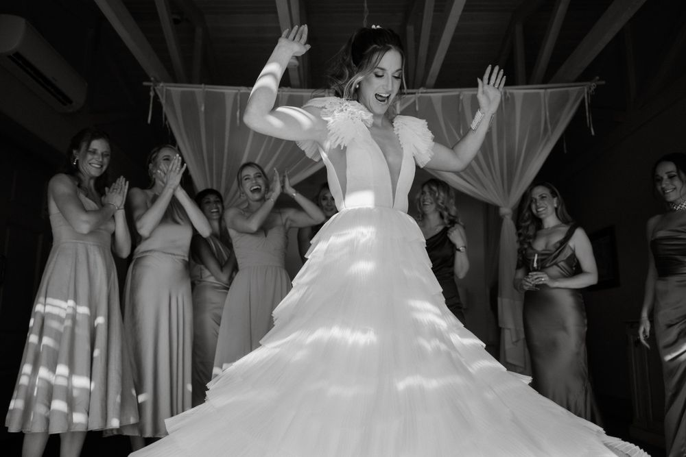 A bride in a layered white wedding dress dances joyfully during a reception with guests in the background.
