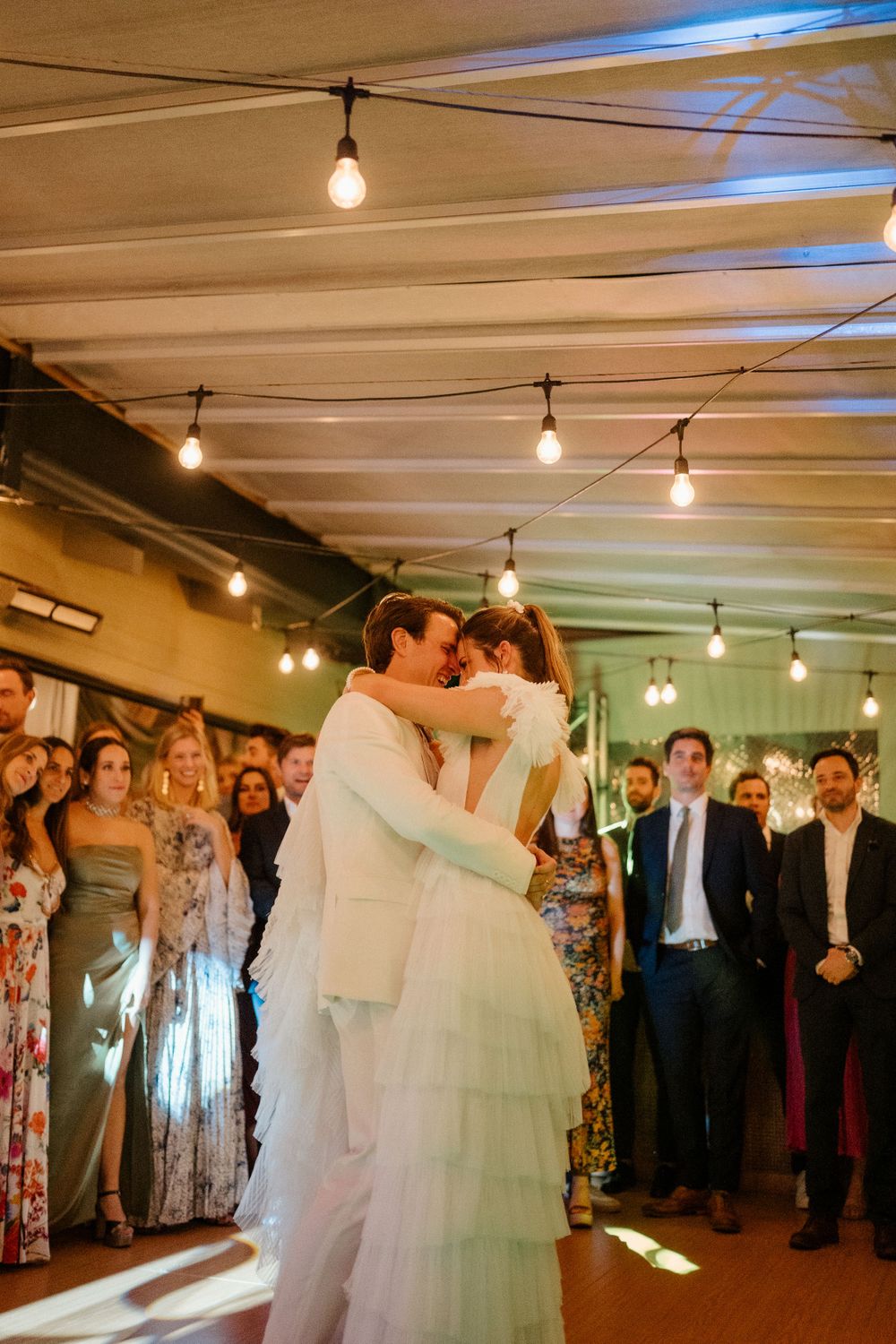 Couple shares first dance surrounded by guests in warmly lit reception venue.