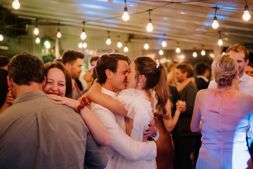 Couples embrace on a crowded dance floor under warm string lights at an indoor reception.