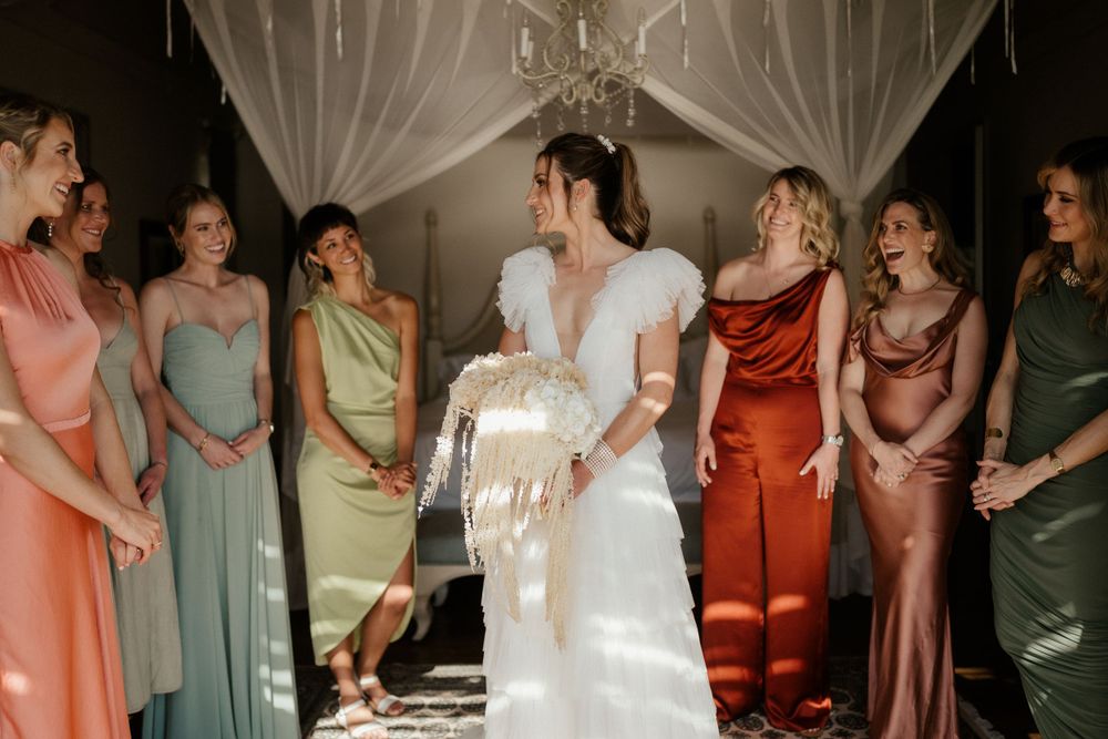 Wedding party in colorful dresses gather around bride in elegant white gown under draped canopy.