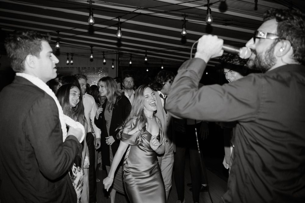 Black and white photo of guests mingling and dancing at an indoor reception with overhead lighting.