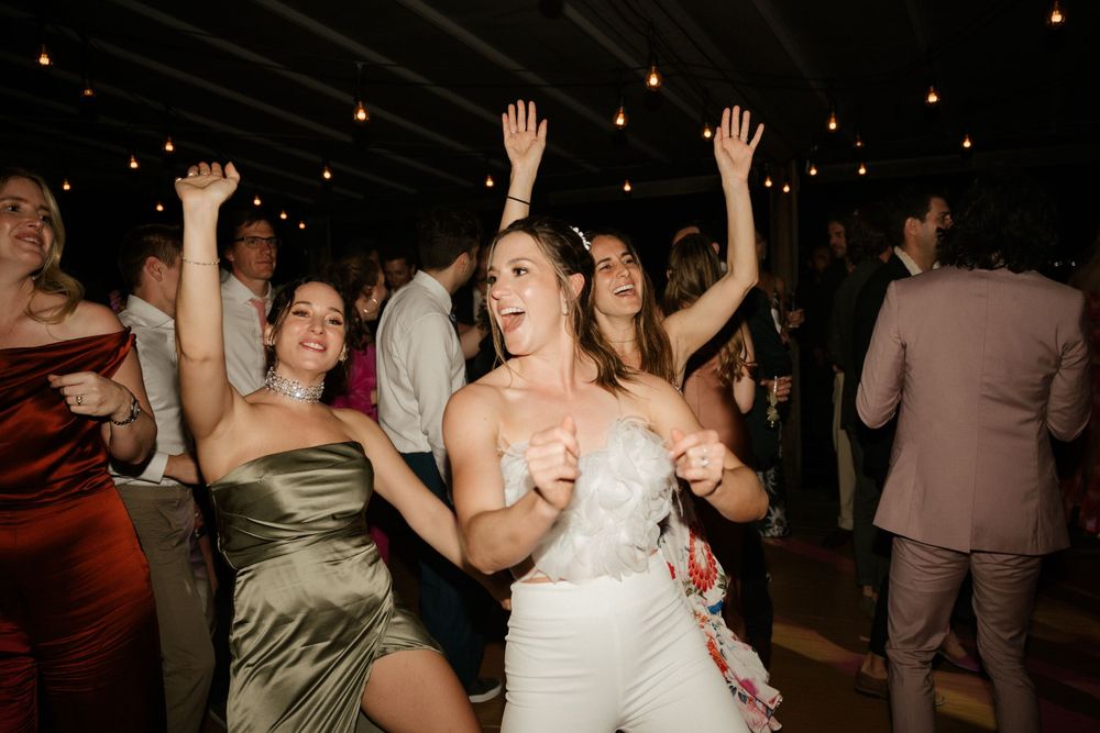 Energetic wedding guests dancing with hands in the air under string lights.