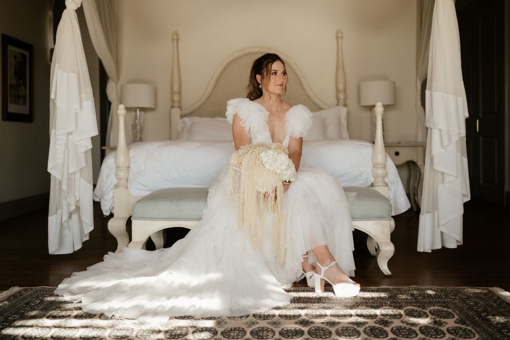 A bride sits elegantly on a white bed in a flowing wedding dress.