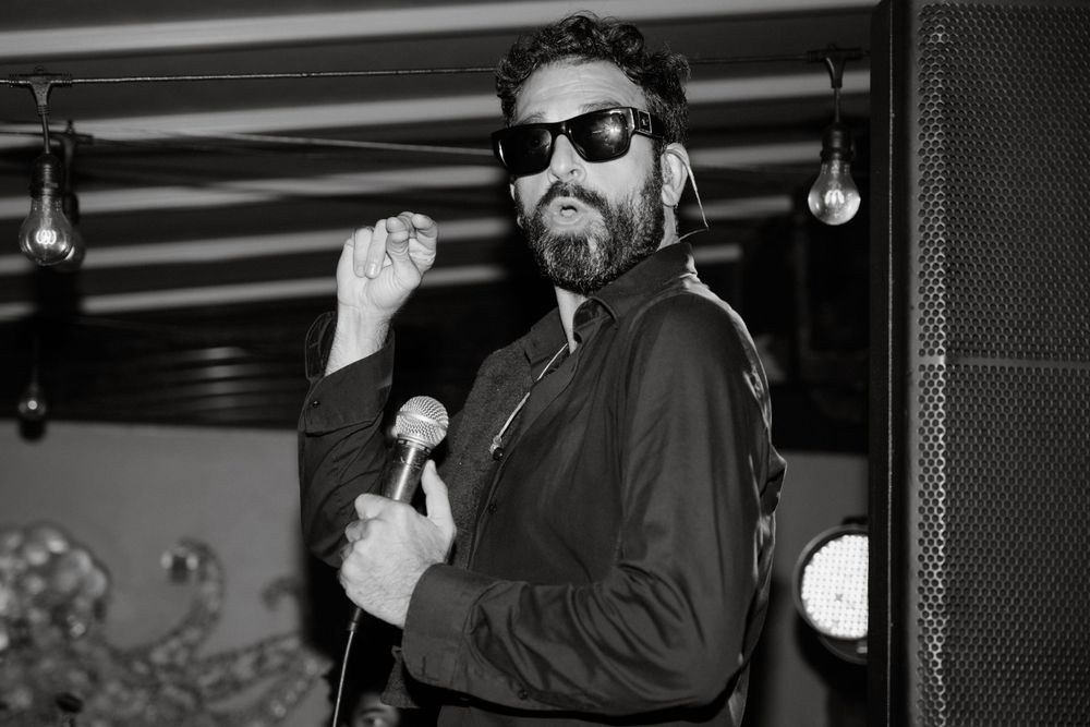Person in sunglasses and dark clothing performing on stage with microphone.