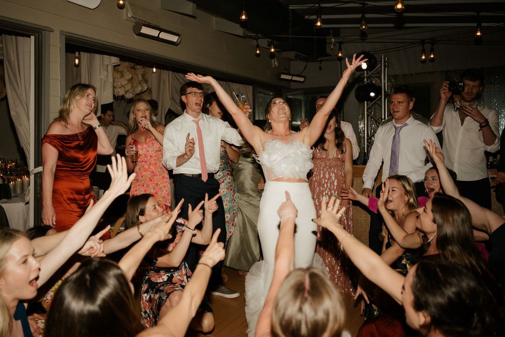 Celebratory dancing scene with guests lifting up someone in white at a festive wedding reception.