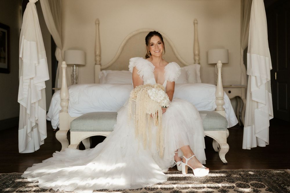 A bride in white dress sits elegantly on a luxurious bed in sunlit room.