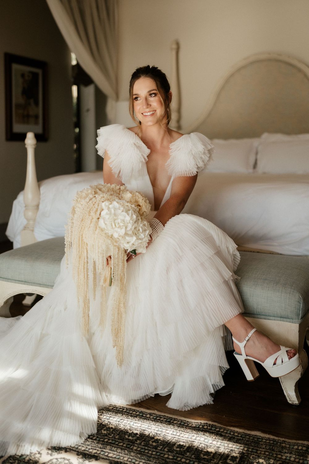 Bride in puffy-sleeved wedding dress sitting with cream-colored bouquet on white bed.