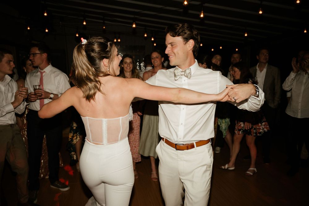 A couple dancing together at a wedding reception in white attire under string lights.