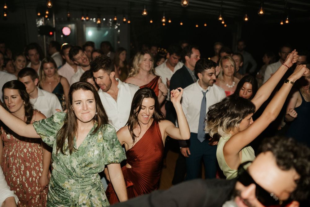 Energetic wedding guests dancing and celebrating on a dimly lit dance floor under string lights.