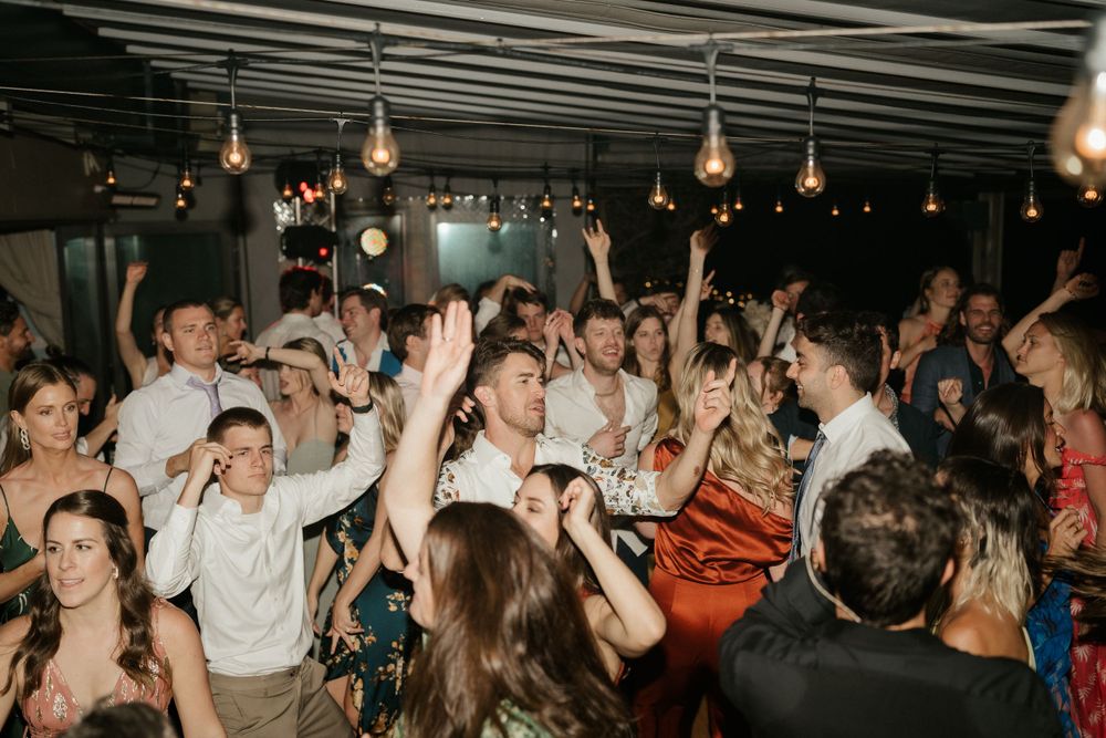 Energetic crowd of wedding guests dancing with raised arms under string lights.