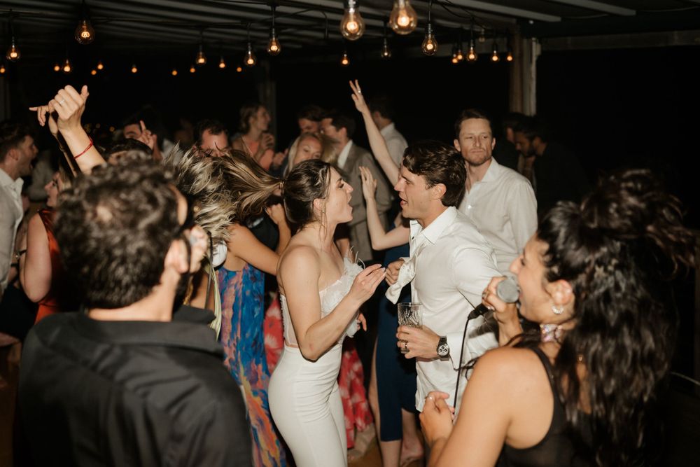 Energetic wedding reception dance party with crowd beneath string lights.
