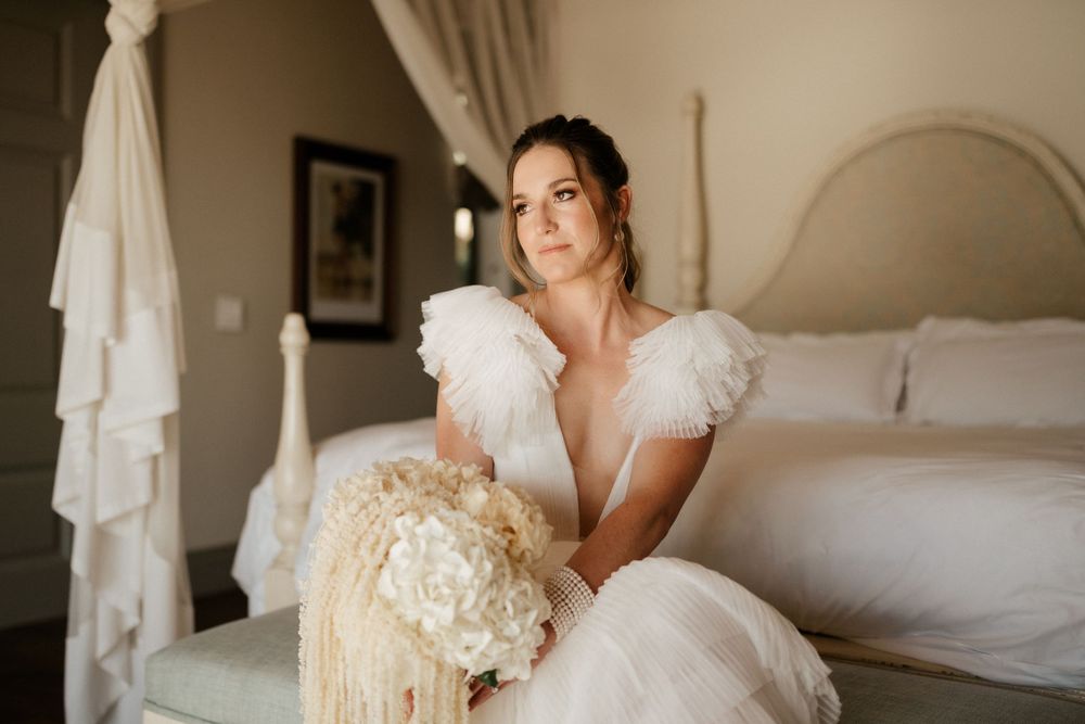 A serene bridal portrait in a white gown with ruffled sleeves sitting on a bed with cream-colored florals.