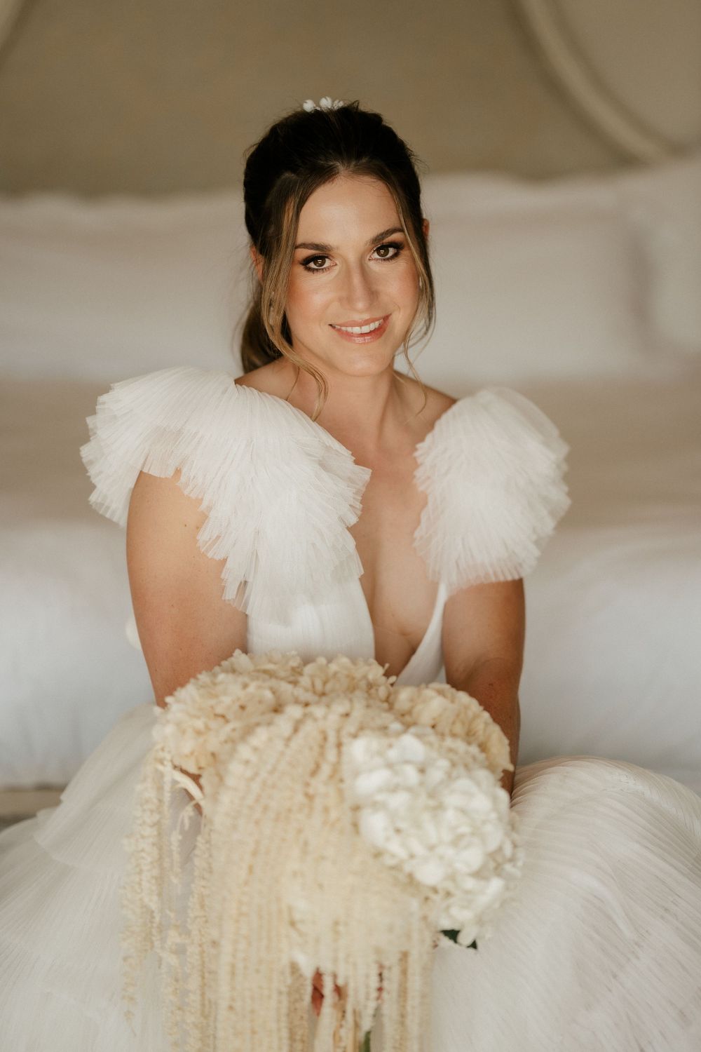 Bride in a white tulle wedding dress sits on a bed holding a bouquet.