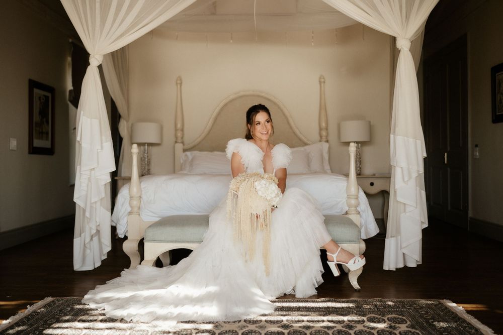 Bride in white dress sits on elegant canopy bed in sunlit room.