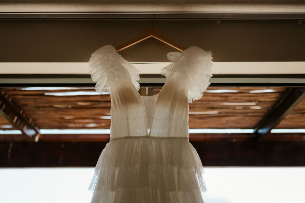 A white wedding dress with feathered cap sleeves hangs from a wooden hanger.