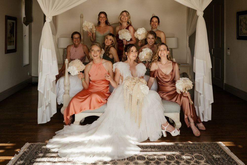 Bridal party in mixed coral and burgundy dresses pose together on white stairs with flowing curtains.