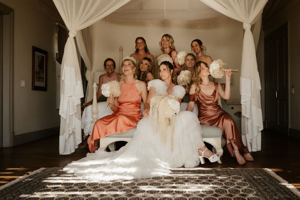 Wedding party gathers in bright room with flowing white curtains and sunlit floor.