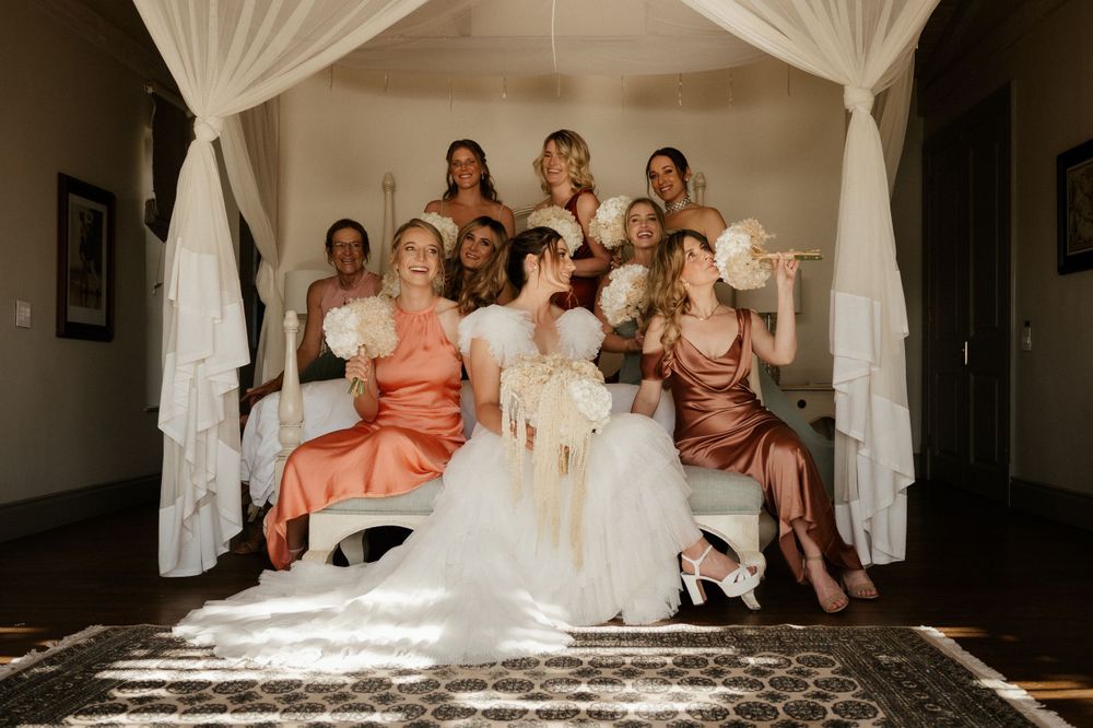 Bridal party poses together on white bedding in a sunlit room.