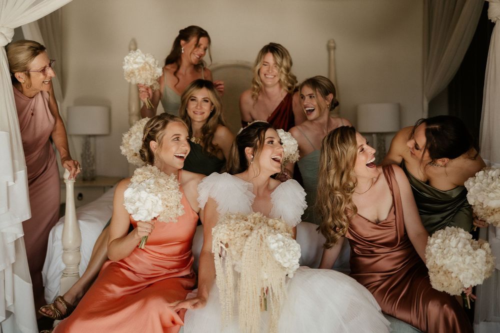 Wedding party with bridesmaids in pink and coral dresses laugh together while holding white bouquets.