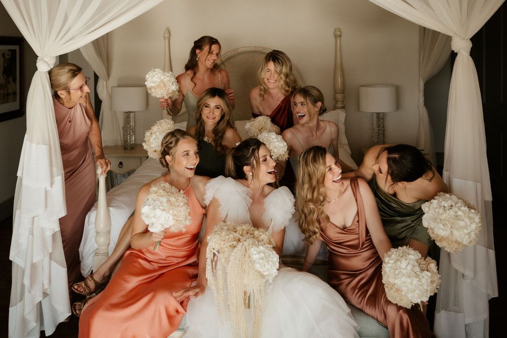 Bridal party in silk dresses shares joyful moment together under flowing white canopy.