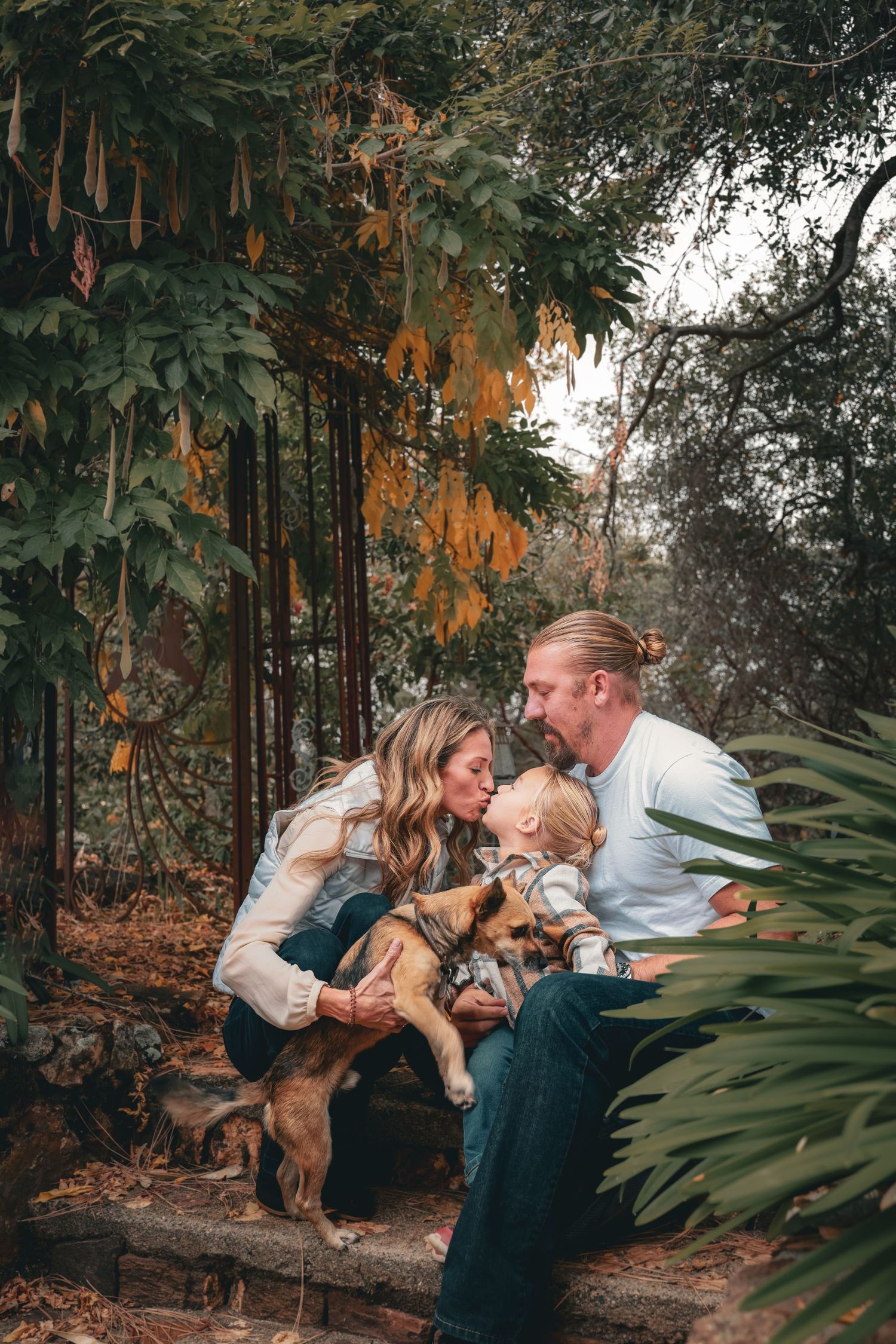 A loving family with a baby and dog share a kiss outdoors in an autumn garden setting.