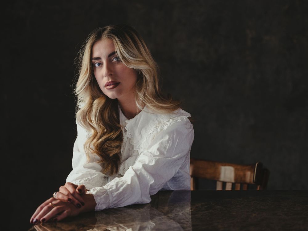 Person in white vintage-style blouse seated at table in dramatic portrait series.