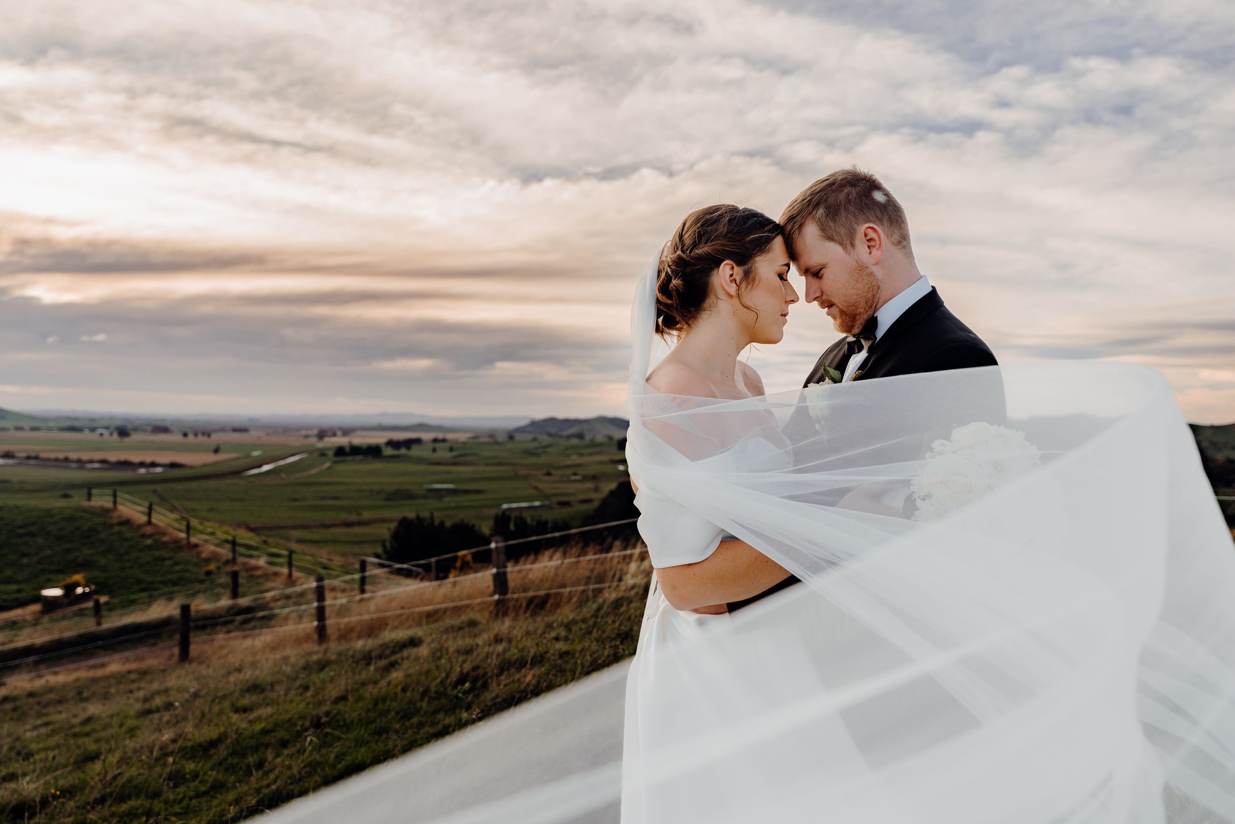 A romantic wedding photo silhouette with flowing veil against a scenic rural landscape at sunset.