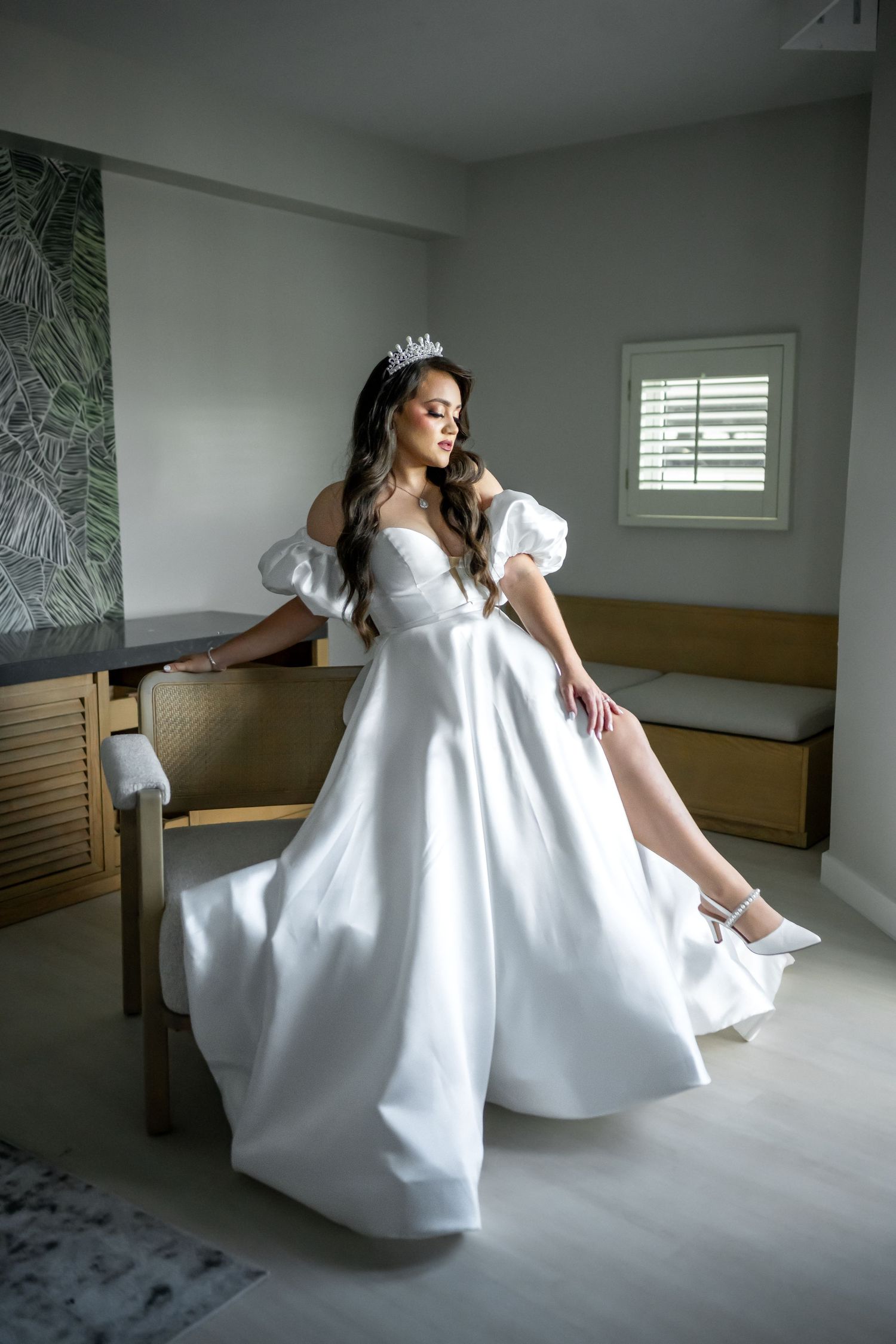 A bride in a flowing white gown poses gracefully near a window in a modern room.