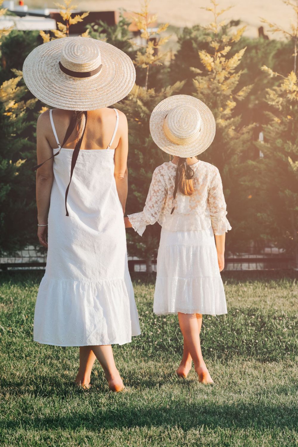 Two figures in white dresses and sun hats stand back-to-back in sunlit garden.