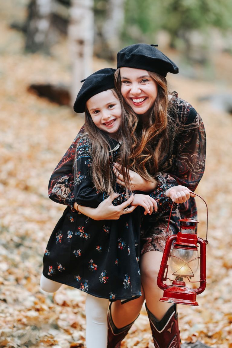 Two people in matching black berets and floral dresses pose with a vintage red lantern among fallen autumn leaves.