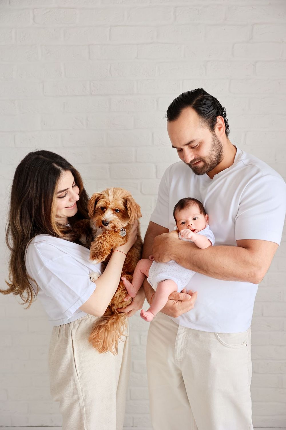 Family portrait with small dog and baby against white brick wall background.